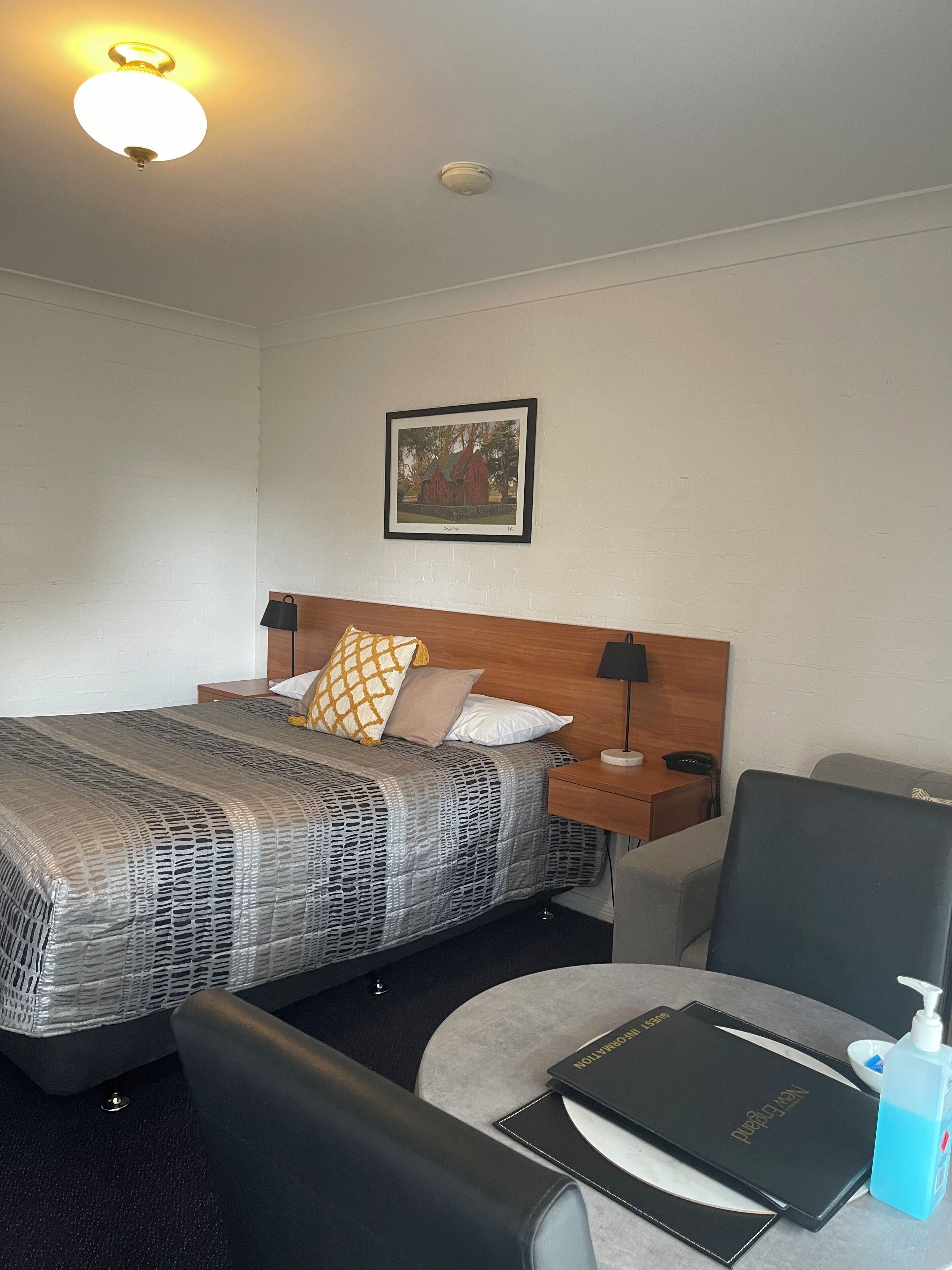 Habitación de hotel con cama, mesitas de noche, una silla, escritorio, lámpara, un cuadro en la pared y una luz en el techo.