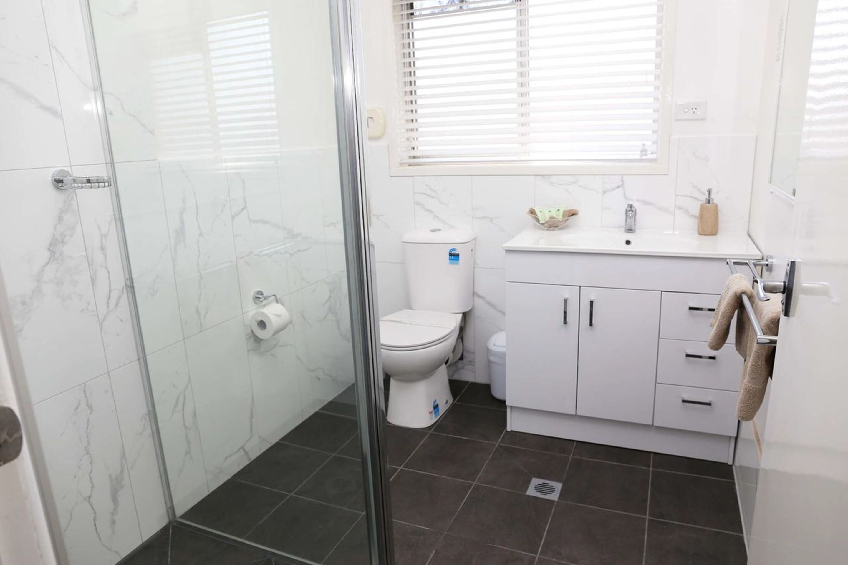 A bathroom with a glass shower enclosure, a toilet, a sink with a mirror, and a window with blinds.