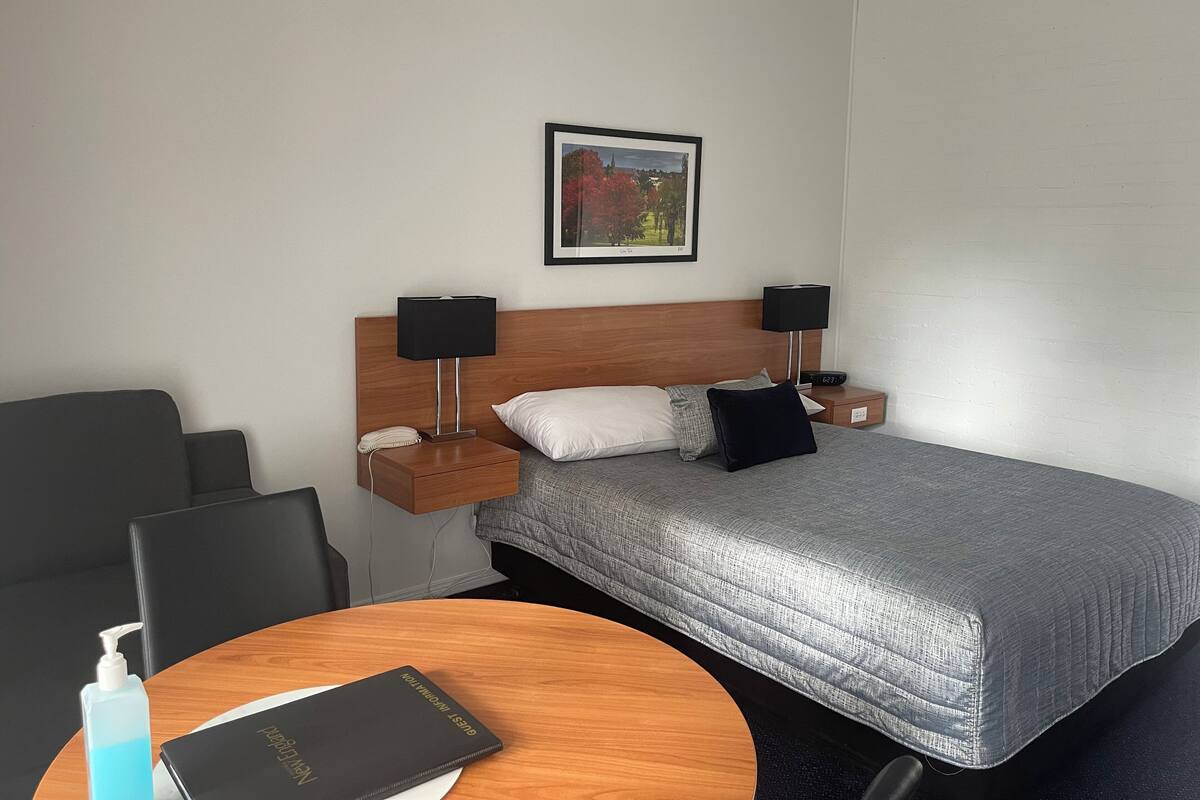 A hotel room with a bed, two bedside tables, a nightstand, a chair, a round table, and a framed picture on the wall.