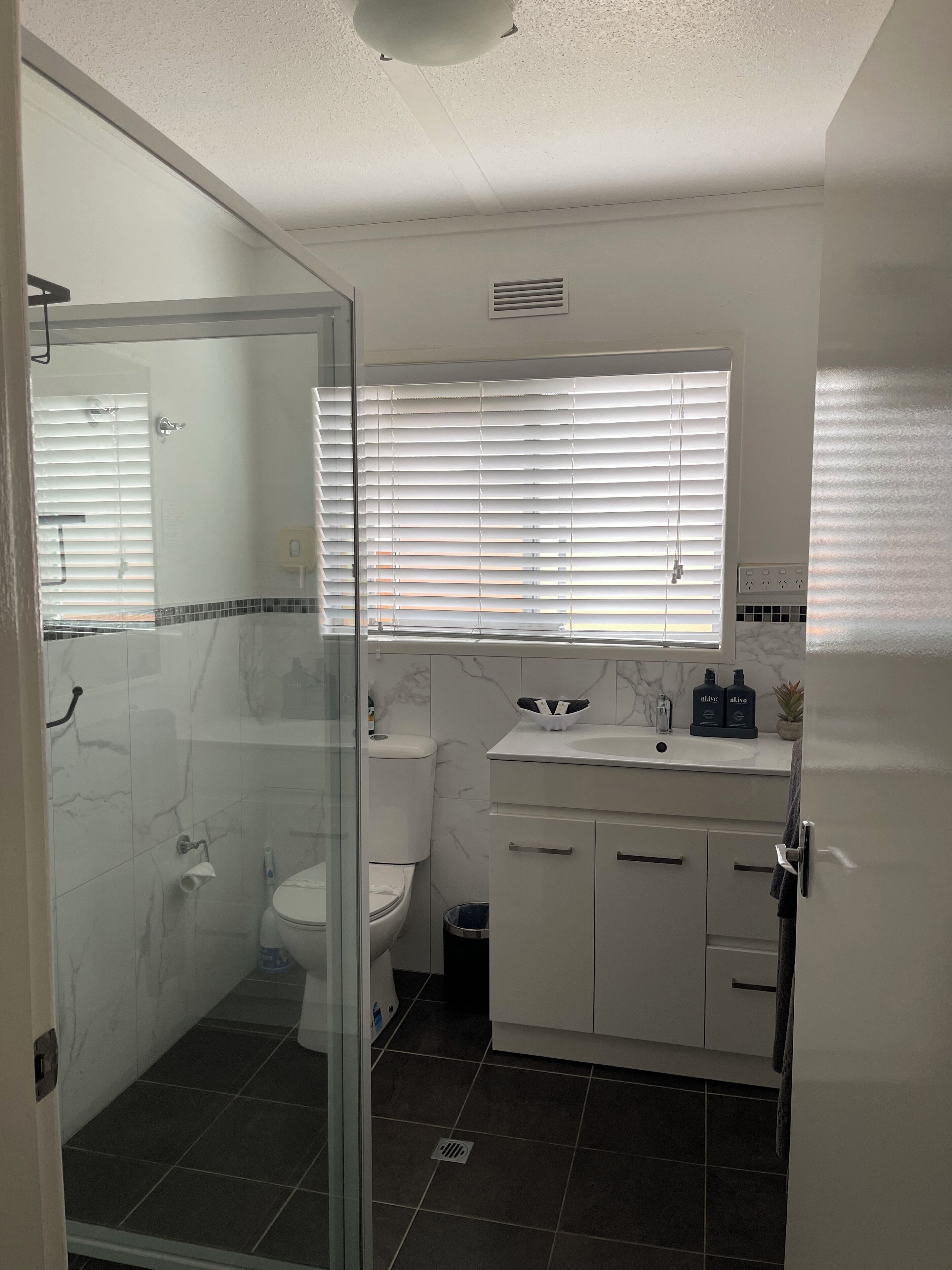 Ein modernes Badezimmer mit einer Duschkabine aus Glas, einer Toilette, einem Waschbecken mit Marmorplatte und einem Fenster mit Jalousien.