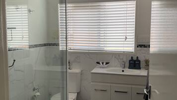 Ein modernes Badezimmer mit einer Duschkabine aus Glas, einer Toilette, einem Waschbecken mit Marmorplatte und einem Fenster mit Jalousien.