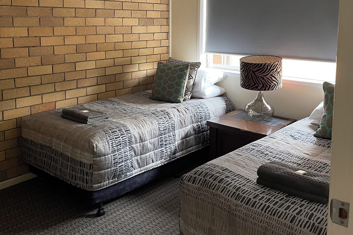 A room with two beds, a brick wall, a window with a blind, and a lamp on a nightstand.