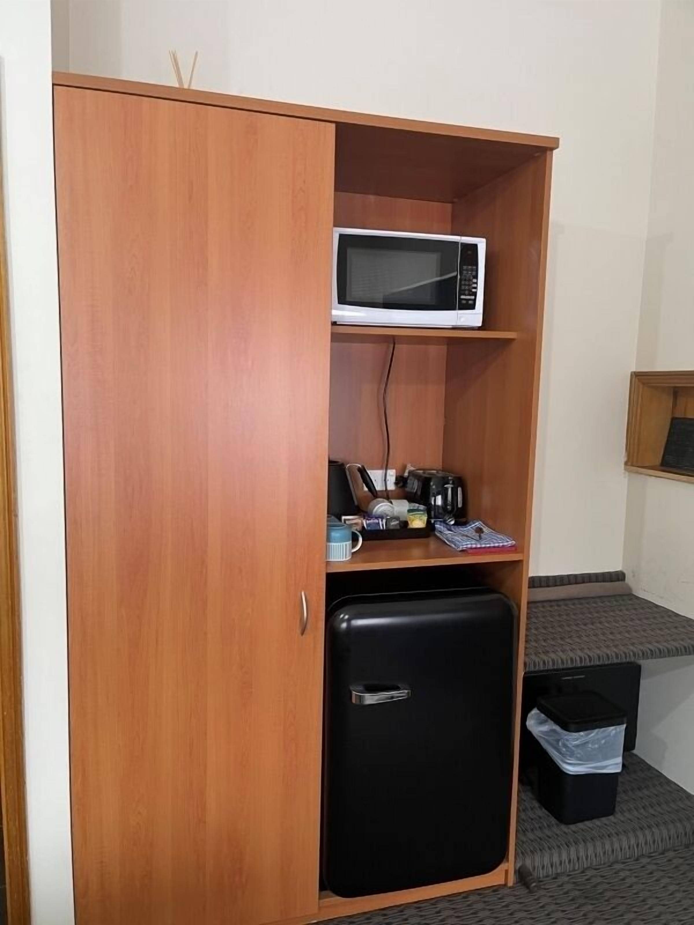 Une pièce de taille modeste, avec une armoire en bois, un réfrigérateur noir, un four à micro-ondes posé sur le dessus, et une petite table sur laquelle se trouvent divers objets.