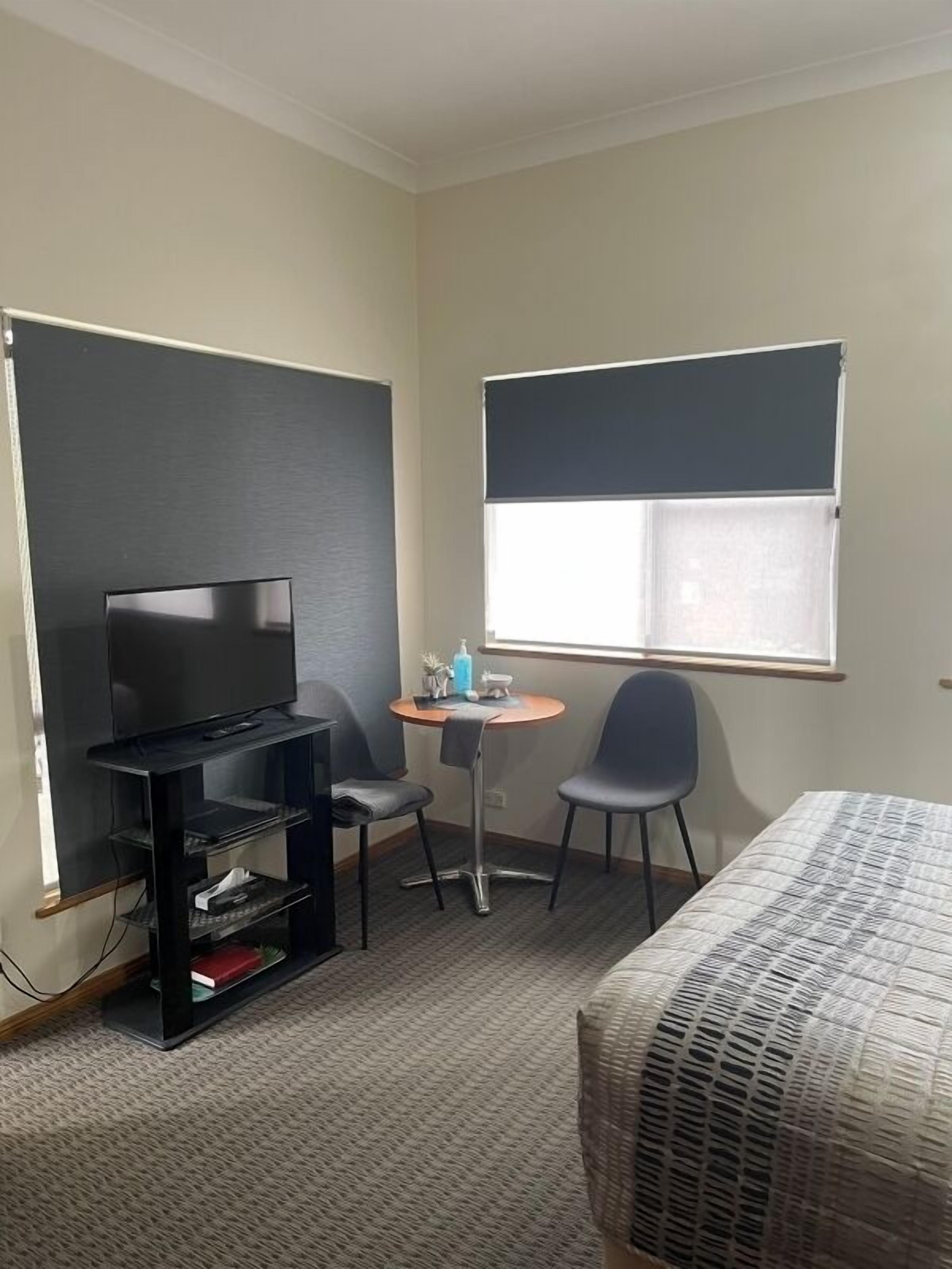 A compact hotel room with a bed, a TV on a stand, a small round table with two chairs, and a window with blinds.