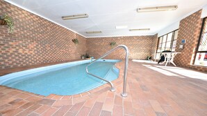 An indoor swimming pool with a brick wall and tiled floor, a metal pool ladder, and a table with chairs in the background. - Horsham Mid City Court Motel (Horsham)