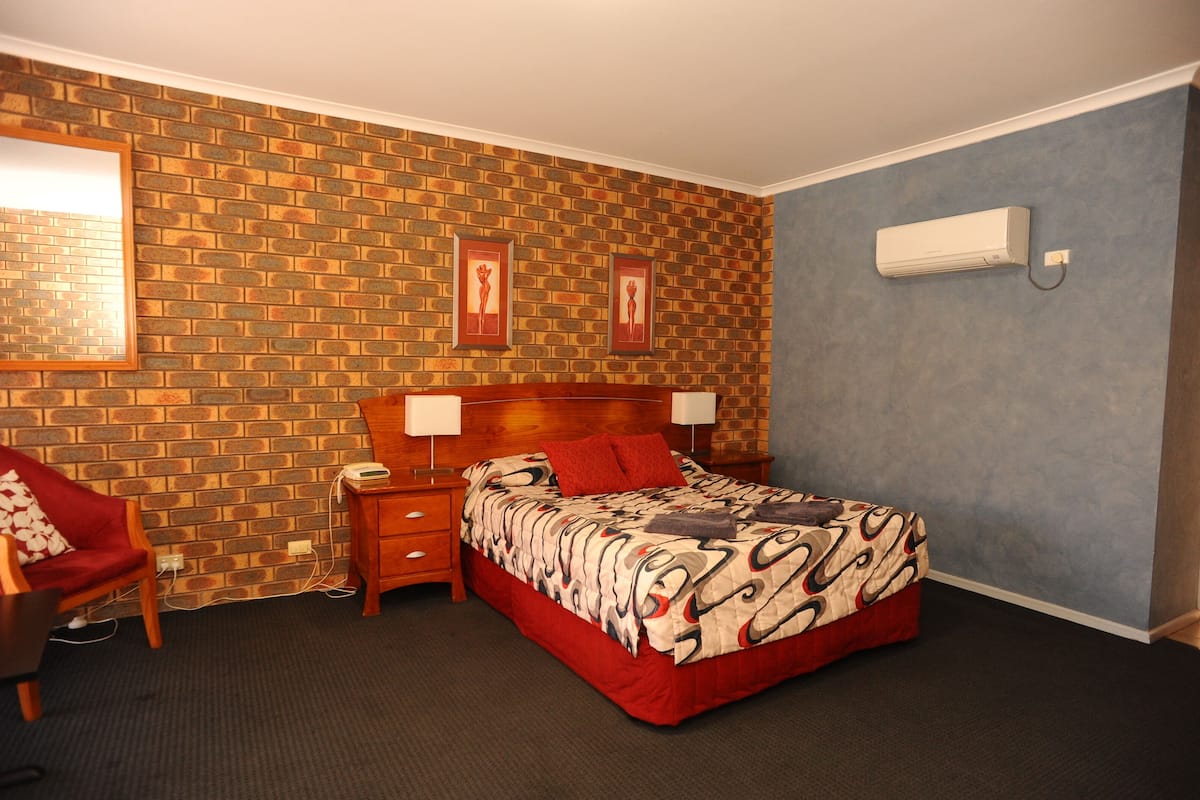Hotellihuone, jossa on tiilikuviollinen seinä, kuviollinen peitto sängyllä, yöpöytä, tuoli ja seinälle asennettu ilmastointilaite.