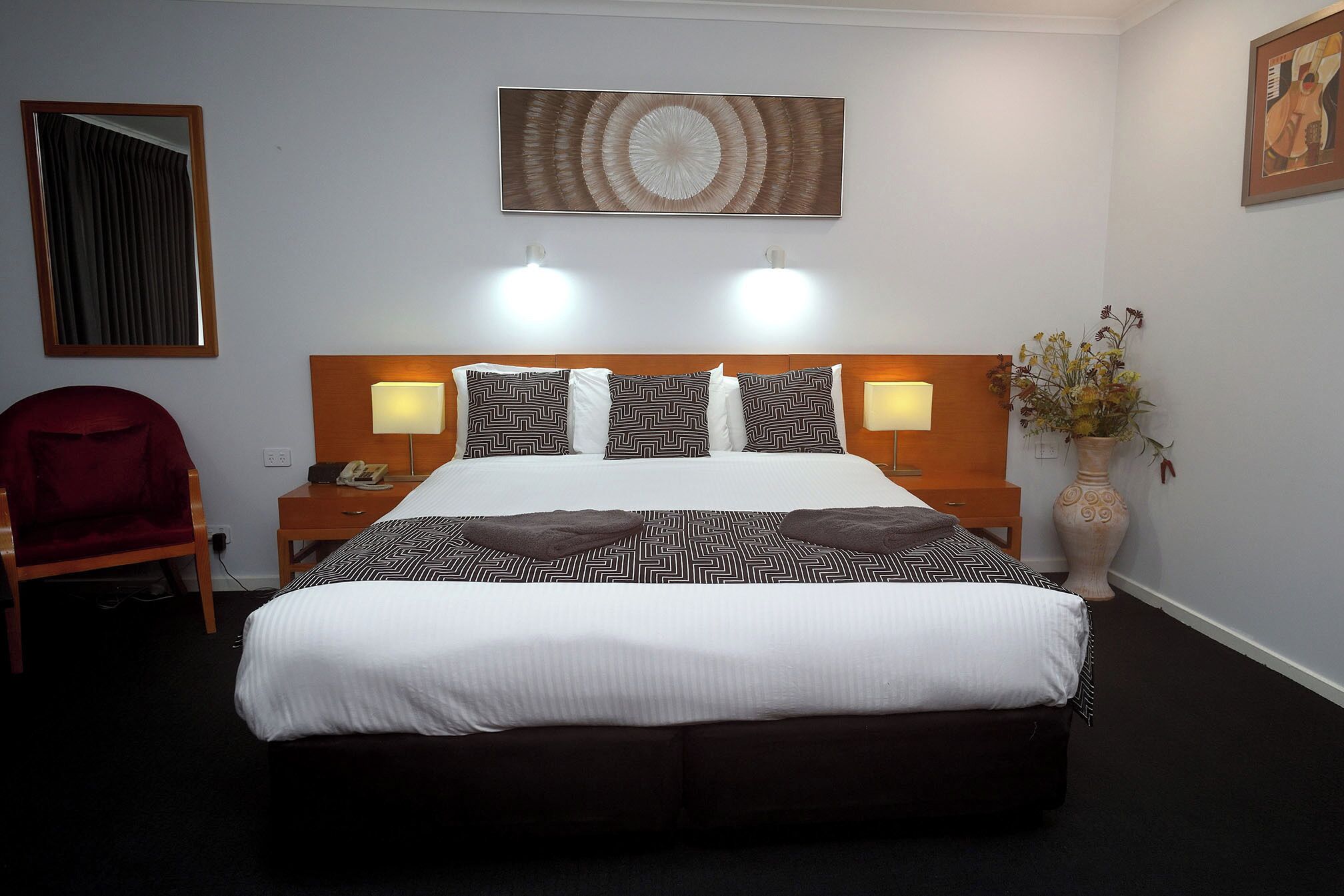 Una habitación de hotel con una cama grande, dos lámparas de noche, un cabecero de madera, una silla roja, un jarrón con flores y un cuadro en la pared.