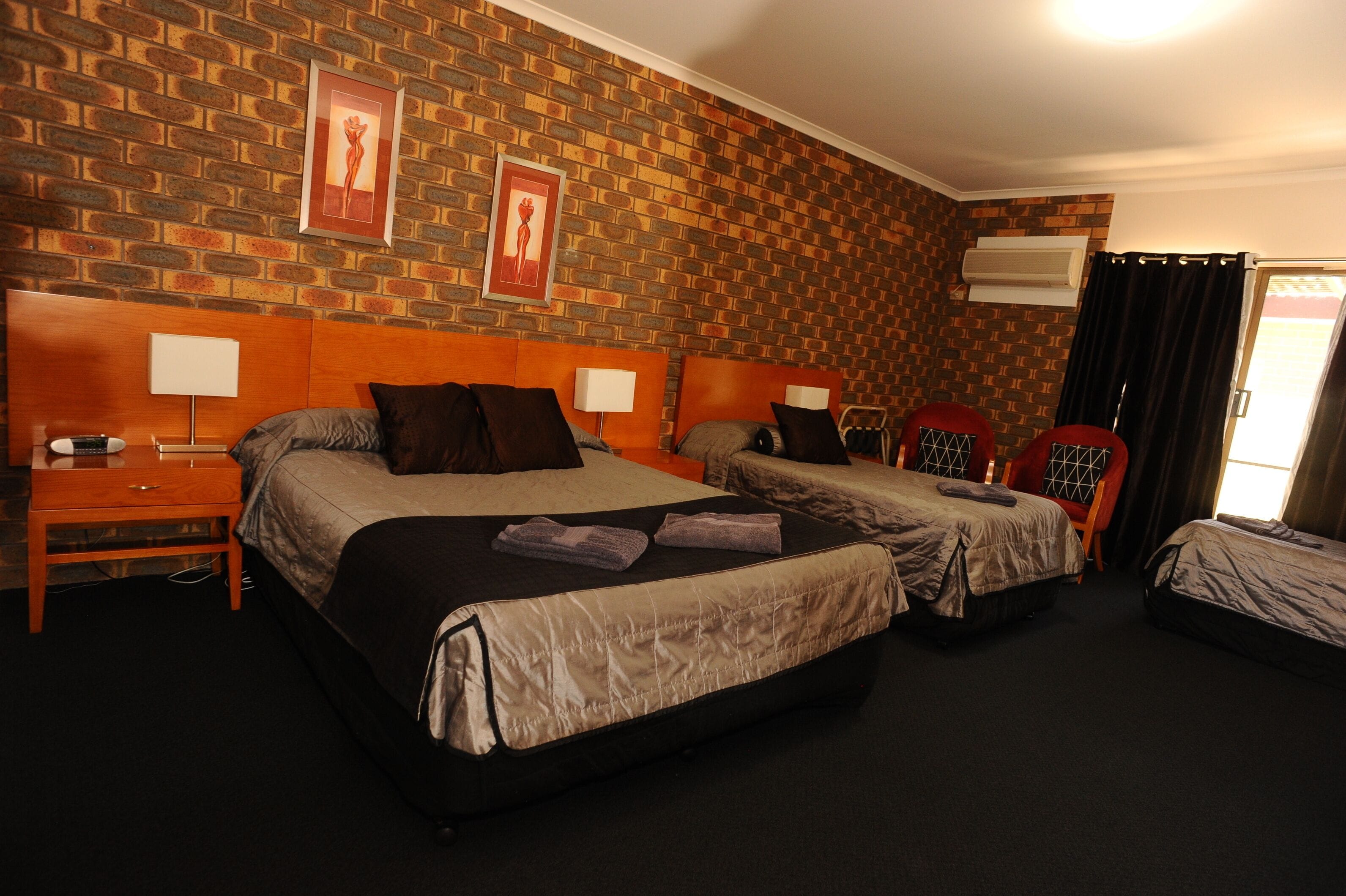 Camera d'albergo con due letti, una parete a mattoni, una testiera in legno, un comodino, una lampada e una finestra con tende.