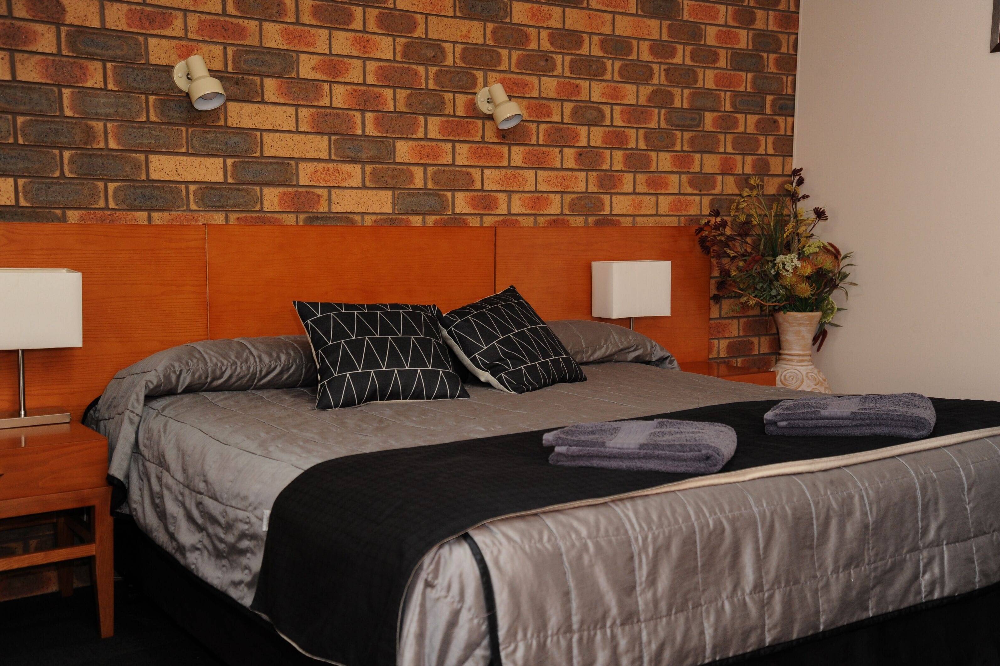 A neatly made bed with a grey and black patterned bedspread, two black pillows, and folded towels. There are two bedside lamps, a wooden nightstand, and a potted plant on the right side.