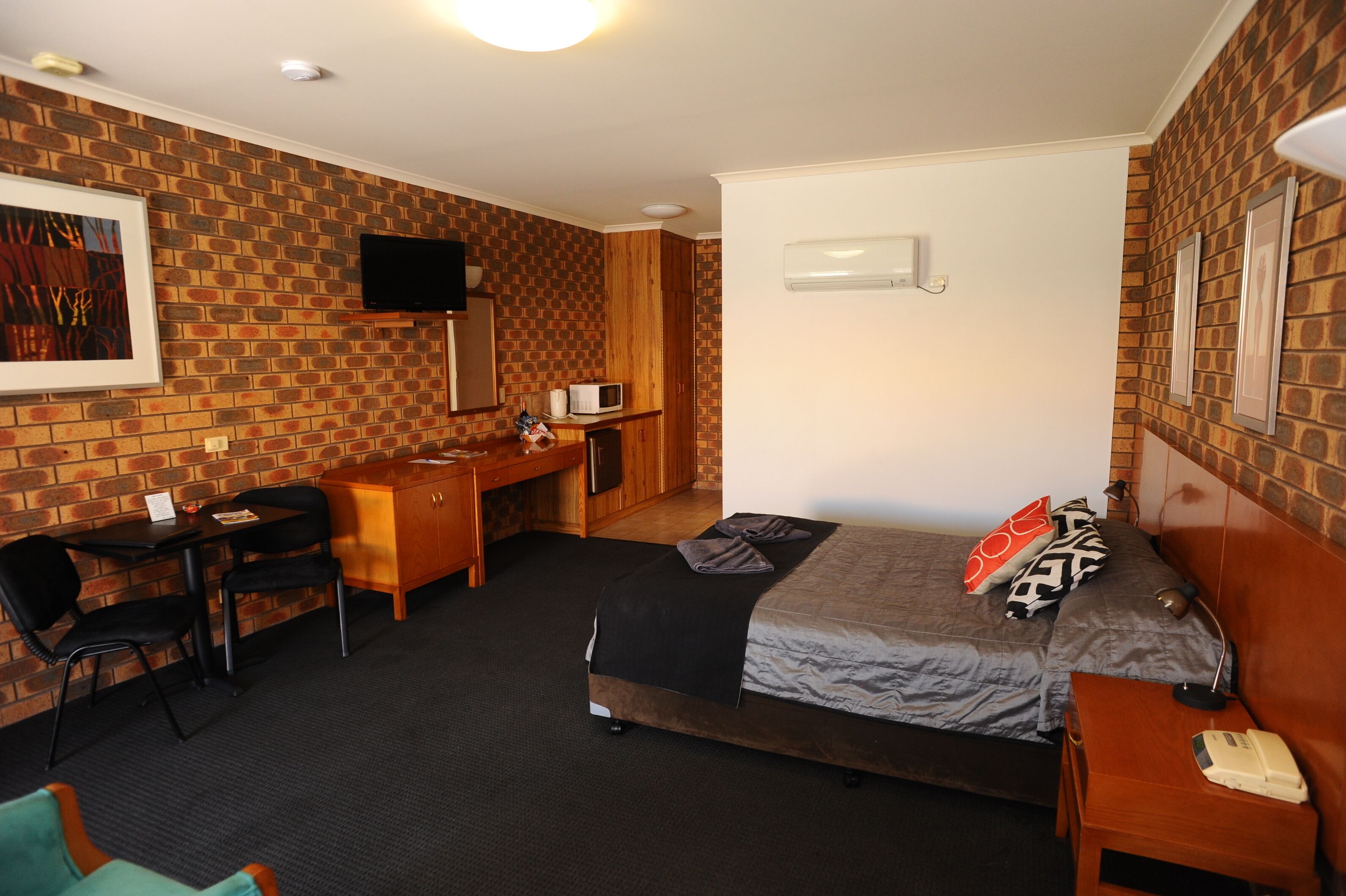 Habitación de hotel con pared de ladrillo, cama con cubrecama gris y negro, escritorio de madera con silla, televisor montado en la pared y mesita de noche con lámpara y teléfono.