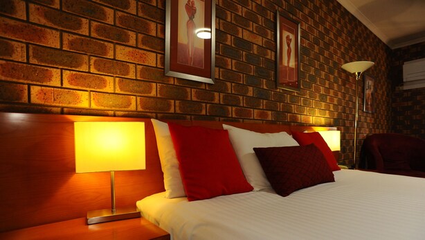 A hotel room with a brick wall, a wooden headboard, a white bed, and red and white pillows. - Horsham Mid City Court Motel (Horsham)