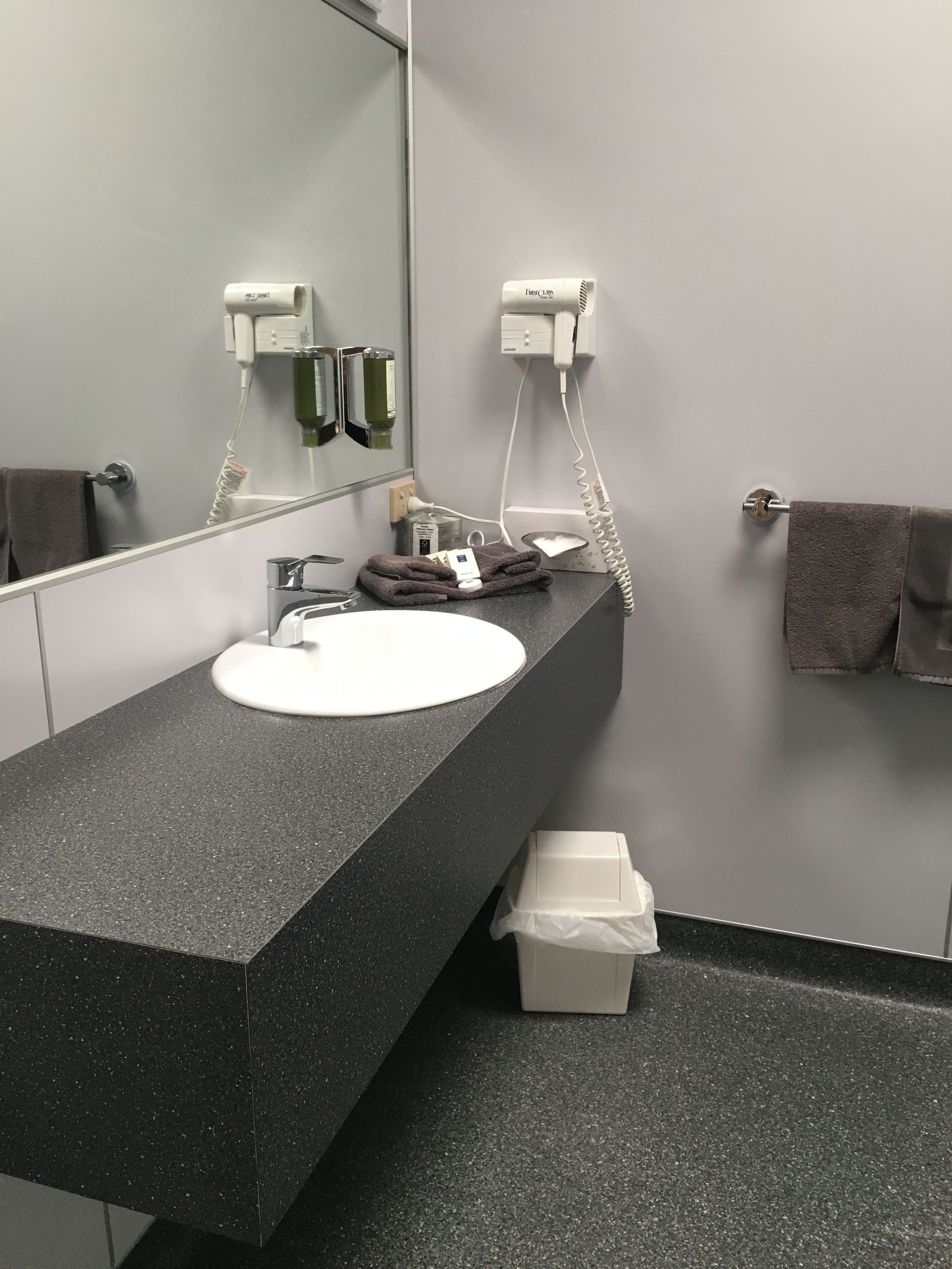 A modern bathroom with a black granite countertop, a white sink, and a large mirror. There are two hair dryers mounted on the wall, a towel rack with a brown towel, and a trash bin.