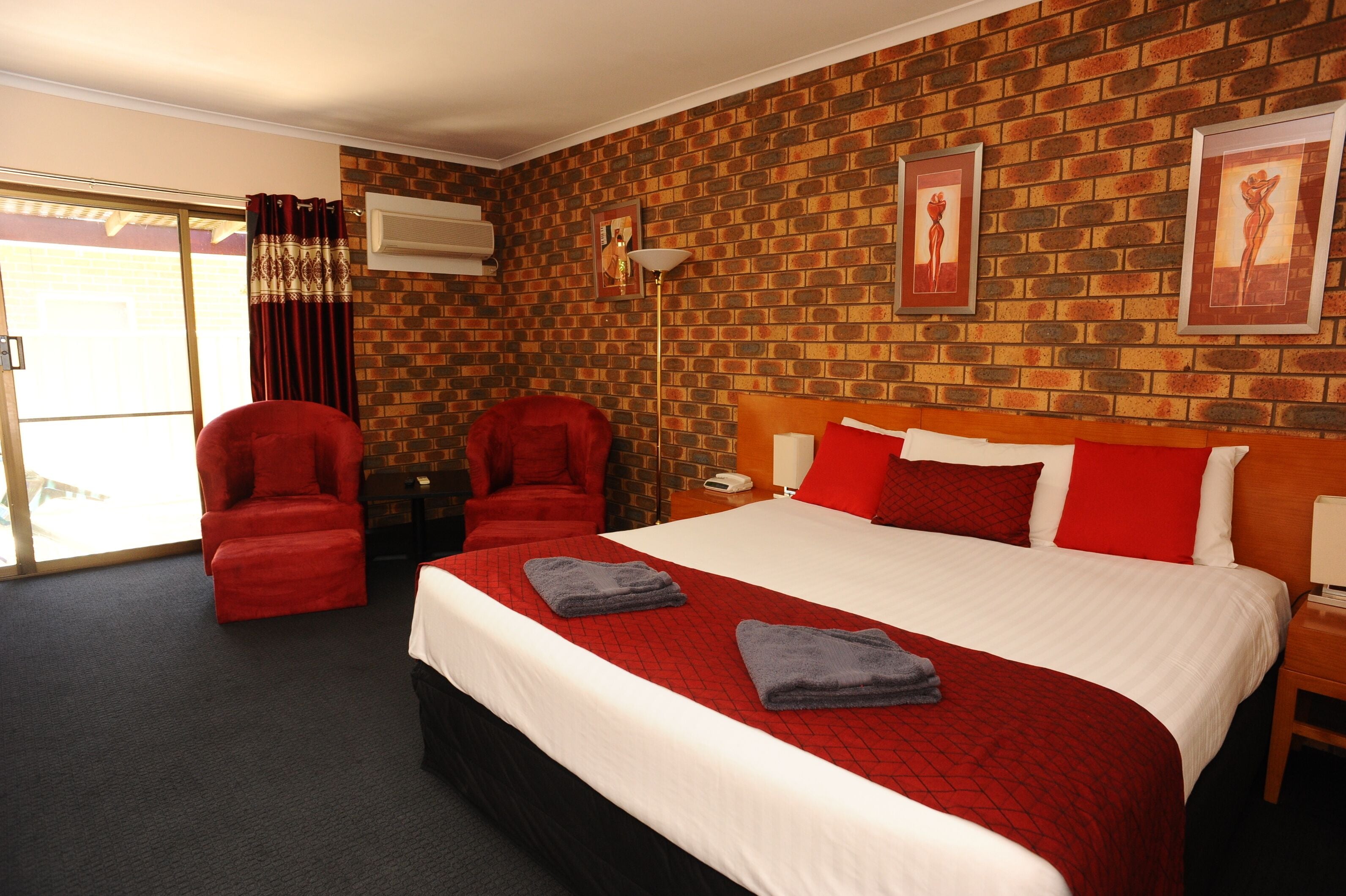 Habitación de hotel con pared de ladrillo, una cama grande con ropa de cama roja y blanca, dos sillones rojos y un gran ventanal corredizo.