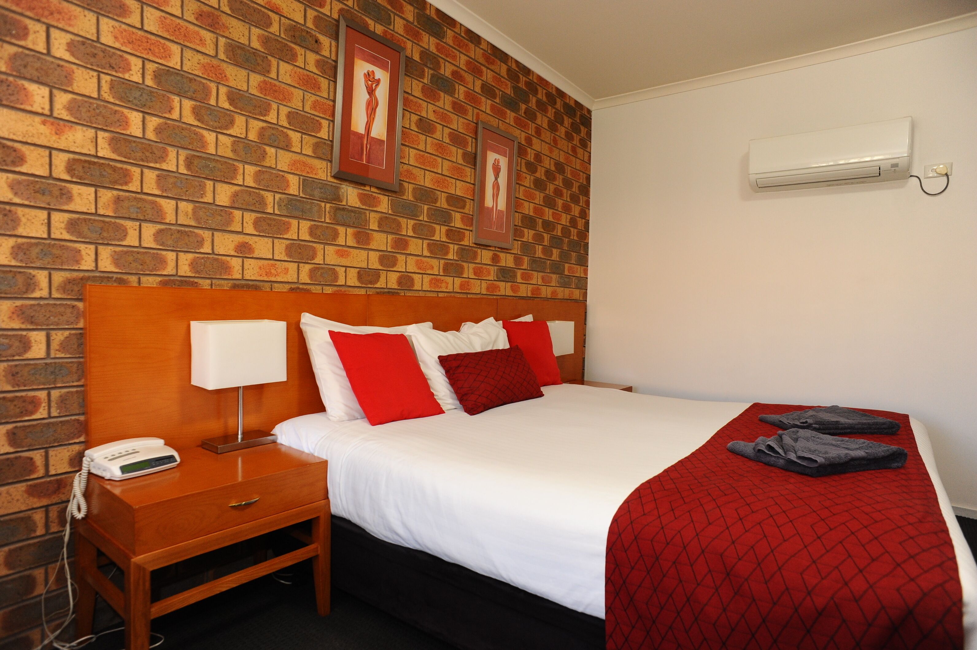 Una habitación de hotel con pared de ladrillo, cabecera de madera, cama blanca con almohadas rojas y blancas, manta roja y aire acondicionado blanco en la pared.