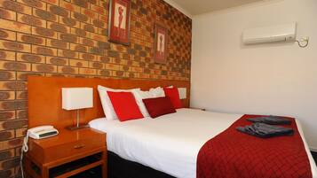 Una habitación de hotel con pared de ladrillo, cabecera de madera, cama blanca con almohadas rojas y blancas, manta roja y aire acondicionado blanco en la pared.