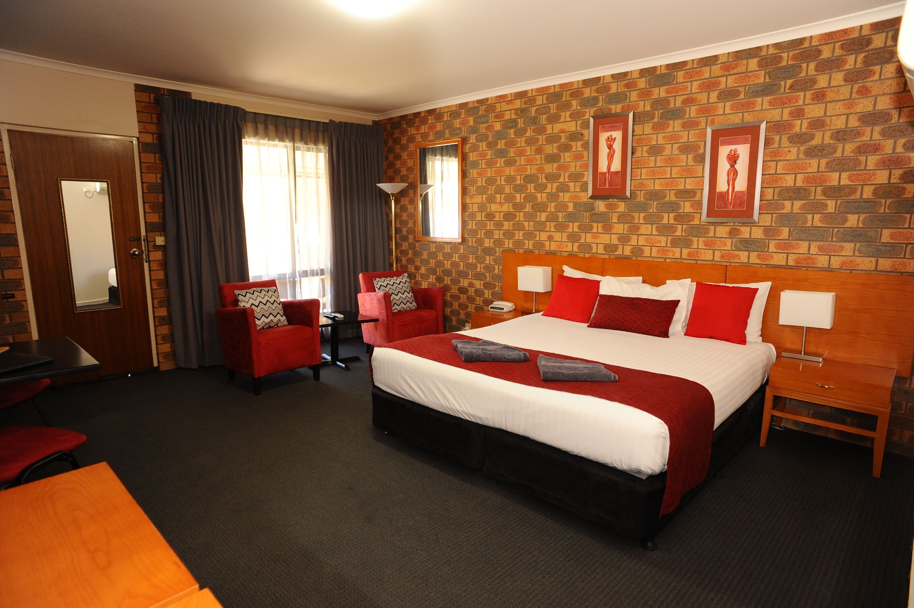 Una habitación de hotel con una cama grande, dos sillones rojos, una mesita de noche de madera, un escritorio y una pared con diseño de ladrillo.