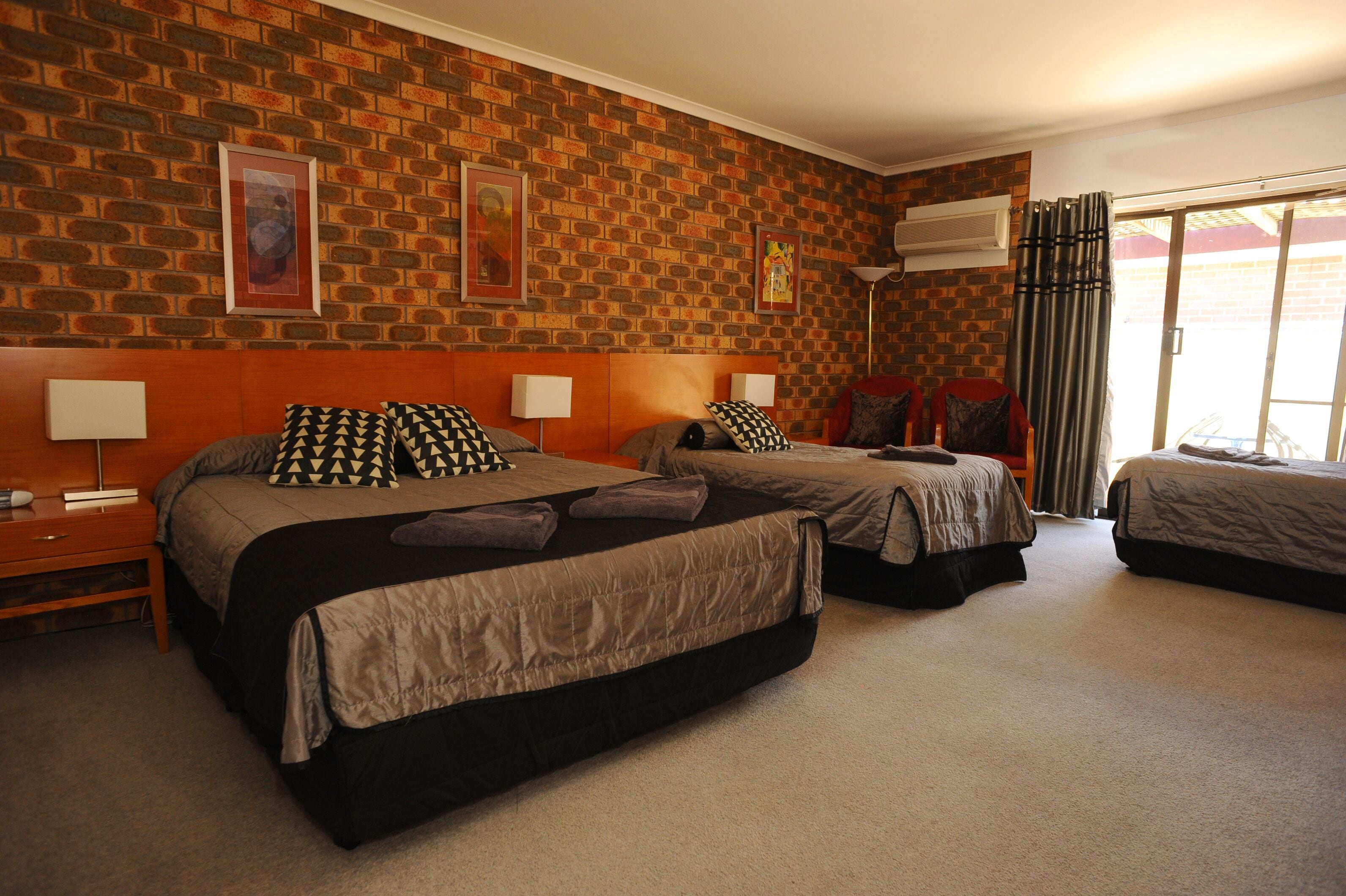 Habitación de hotel con tres camas, pared de ladrillo y un ventanal con cortinas.
