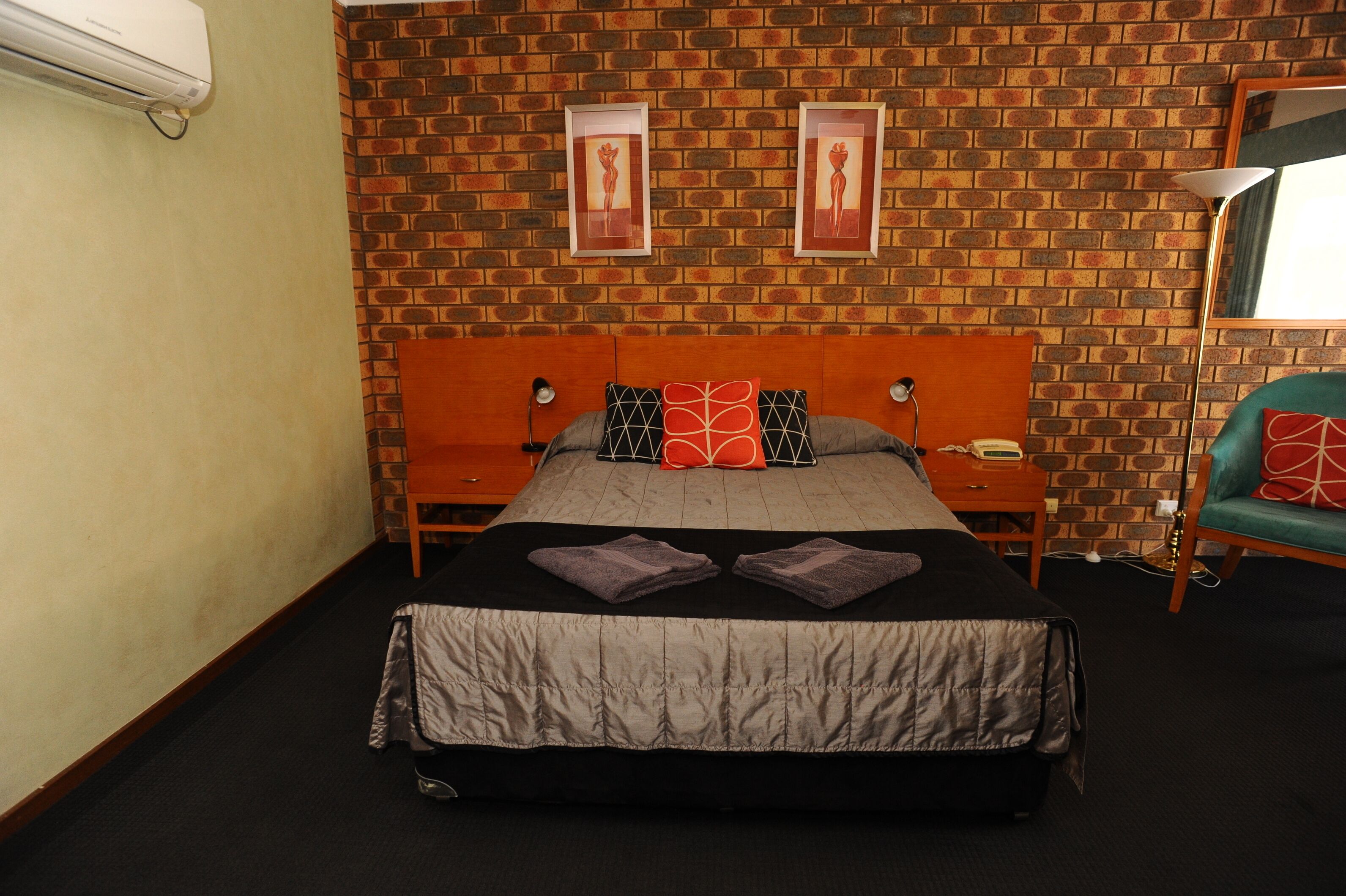Habitación de hotel con pared de ladrillo, cama con colcha gris y negra, dos almohadas, mesita de noche con lámpara y sillón con cojín rojo.