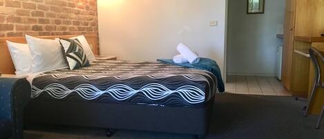 Quarto de hotel com parede de tijolos, cama com roupa de cama estampada, mesa de cabeceira e ar condicionado fixado na parede.