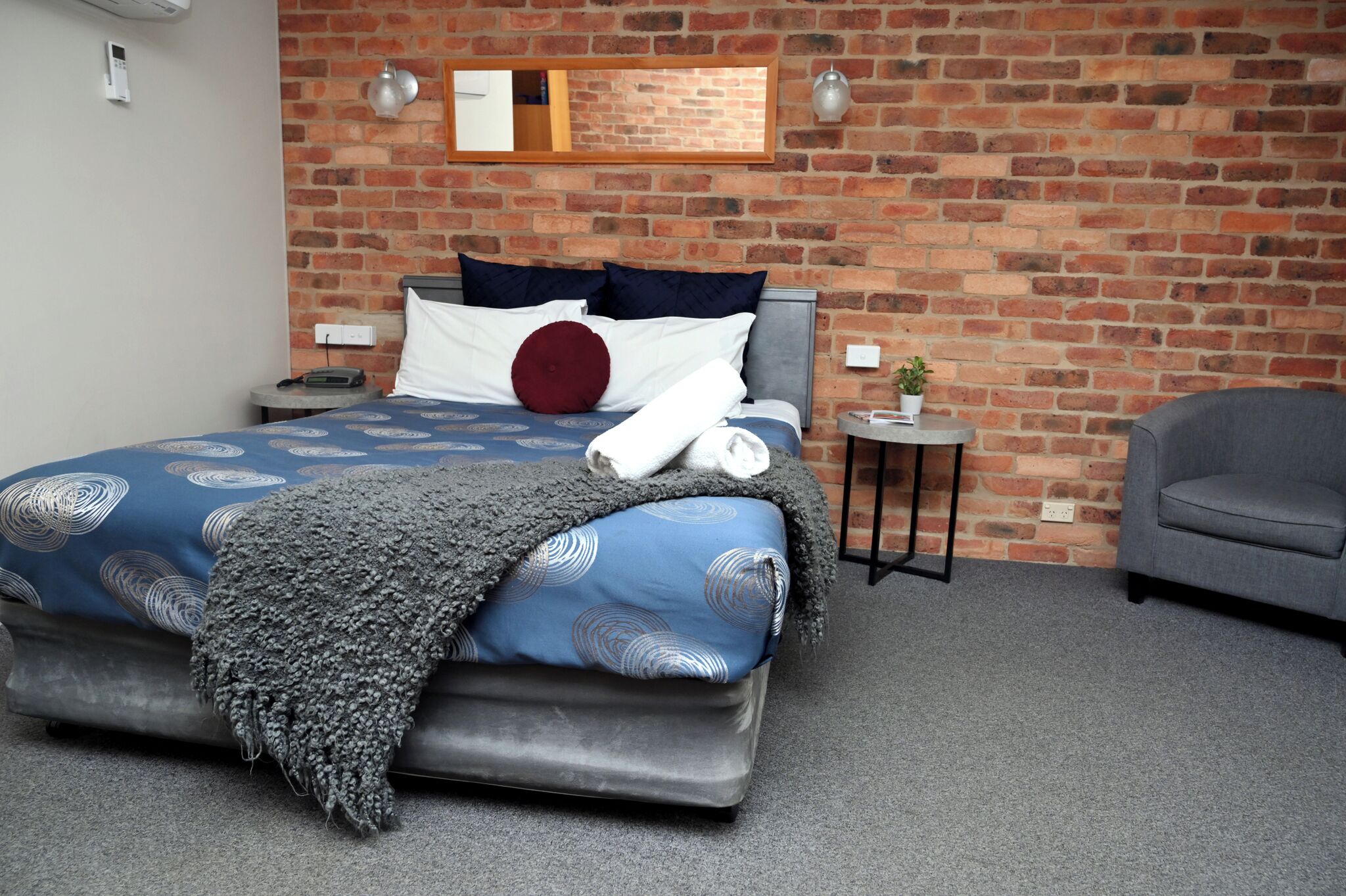 A hotel room with a brick wall, a bed with a patterned bedspread, a grey armchair, and a small side table with a plant.