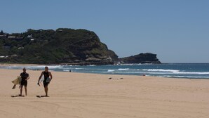 Una playa con dos personas cargando tablas de surf, un acantilado rocoso y un cielo despejado.