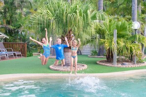 A pool area with a circular water feature, surrounded by palm trees and greenery. - The Palms At Avoca (Avoca Beach)
