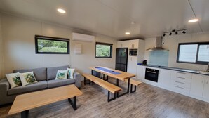 Een ruimtelijk ingerichte leefruimte met een bank, twee houten tafels, een kitchenette en een raam met uitzicht op groen.