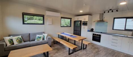Un espacio habitable compacto con sofá, dos mesas de madera, kitchenette y ventana con vistas a zonas verdes.