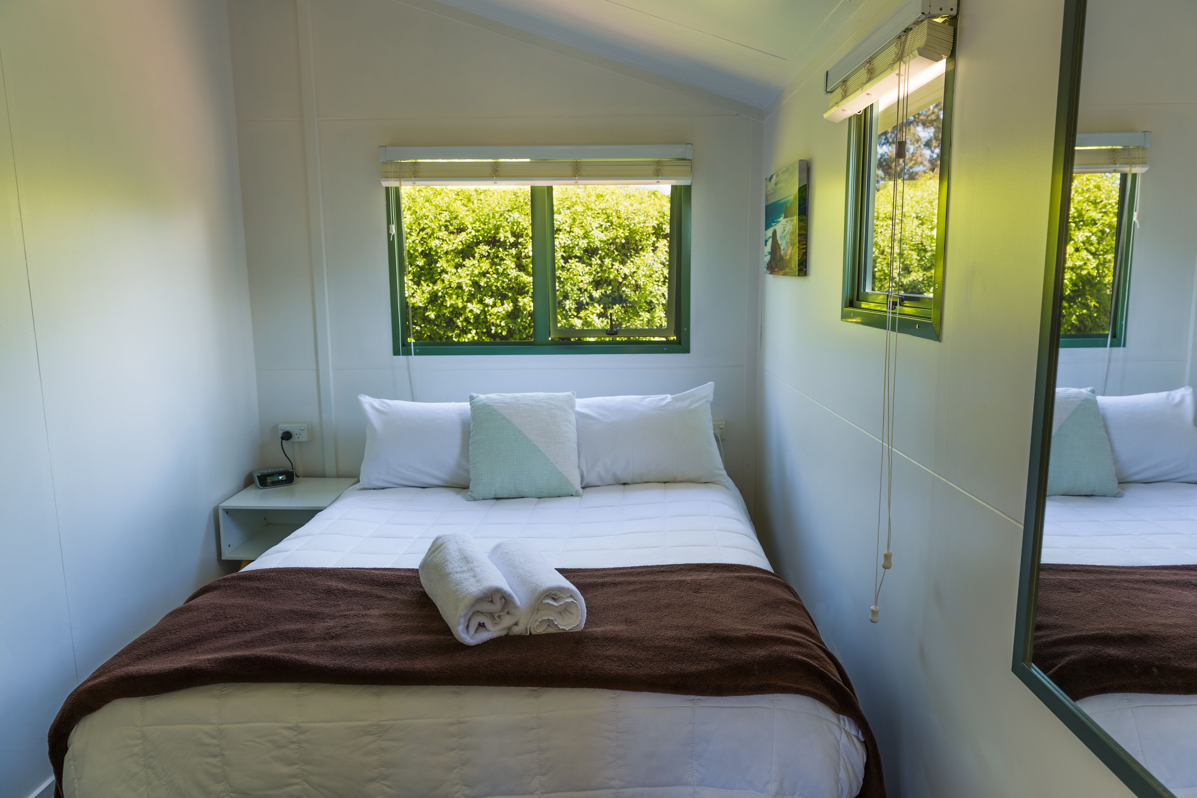 Ein ordentlich bezogenes Bett mit einer braunen Decke, weißen Kissen und einem Paar gefalteter Handtücher daneben. Ein Fenster mit grünen Rollos und Blick auf Bäume.