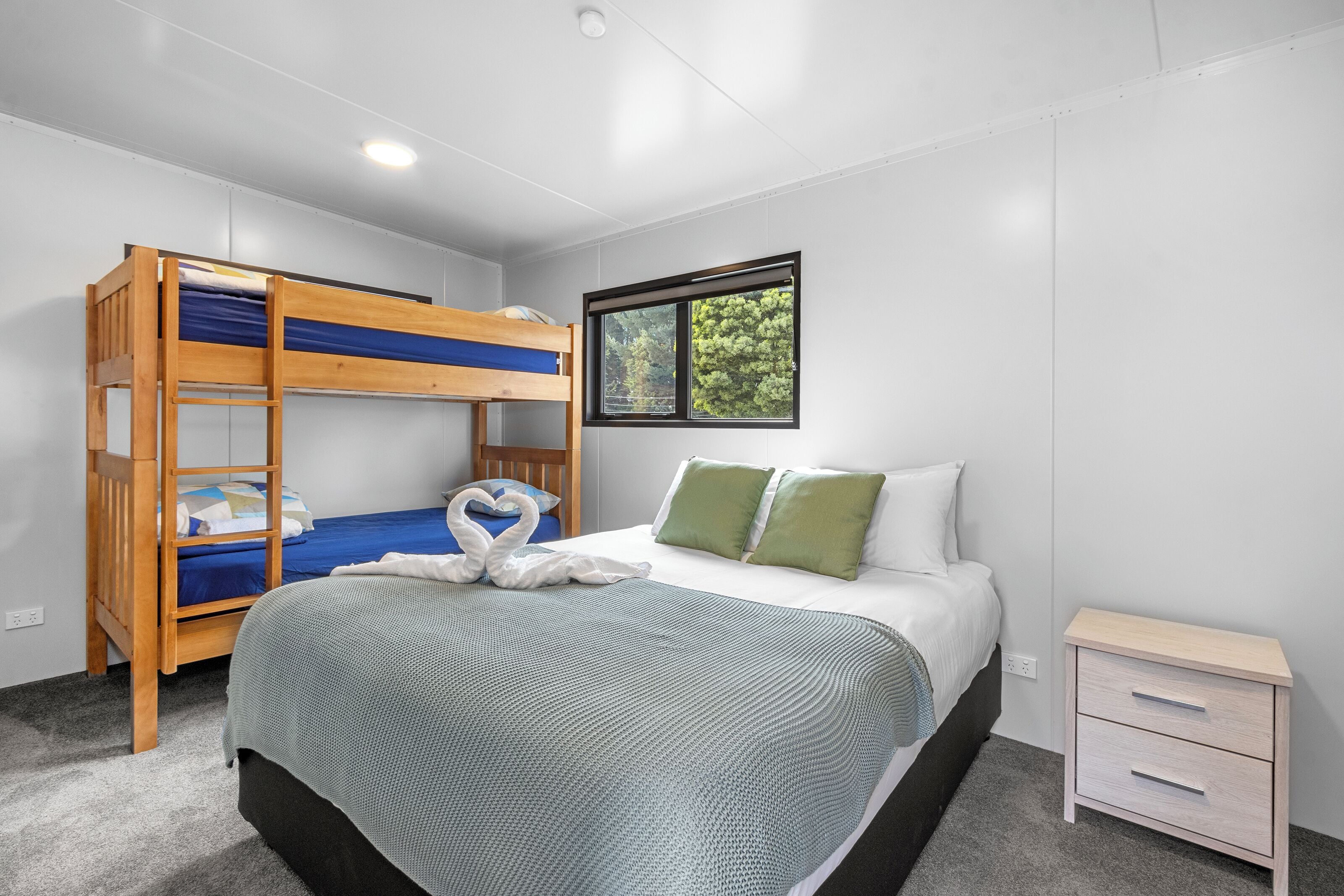 Una habitación con una cama litera, una cama individual con almohadas verdes, una mesita de noche y una ventana con vistas a árboles.