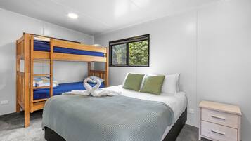 Una habitación con una cama litera, una cama individual con almohadas verdes, una mesita de noche y una ventana con vistas a árboles.