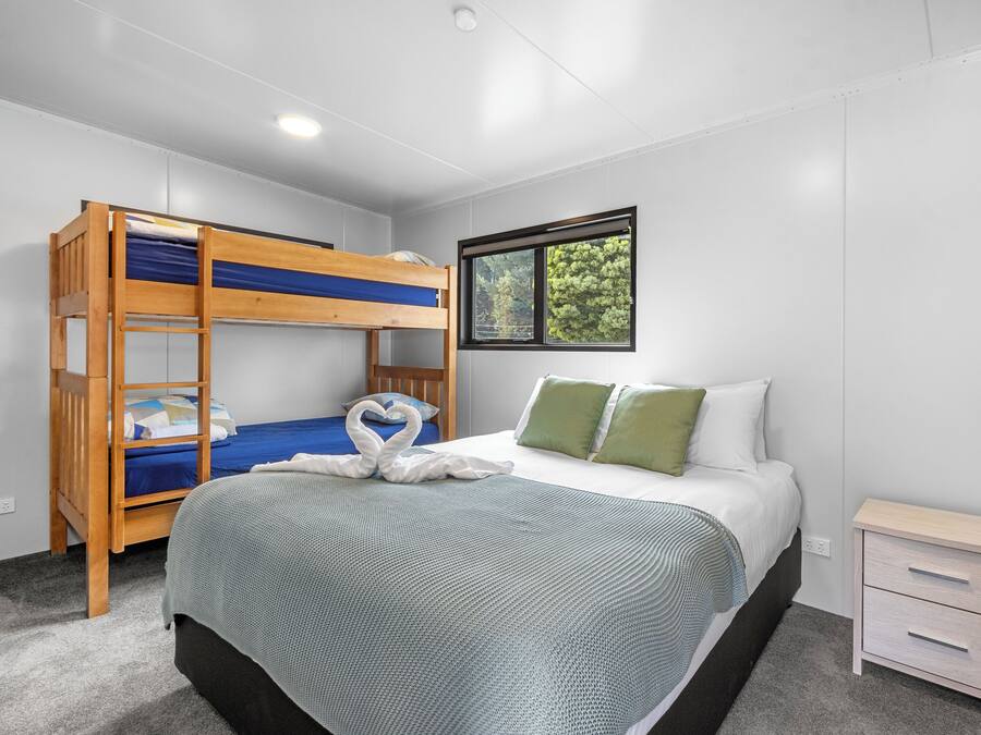 Una habitación con una cama litera, una cama individual con almohadas verdes, una mesita de noche y una ventana con vistas a árboles.