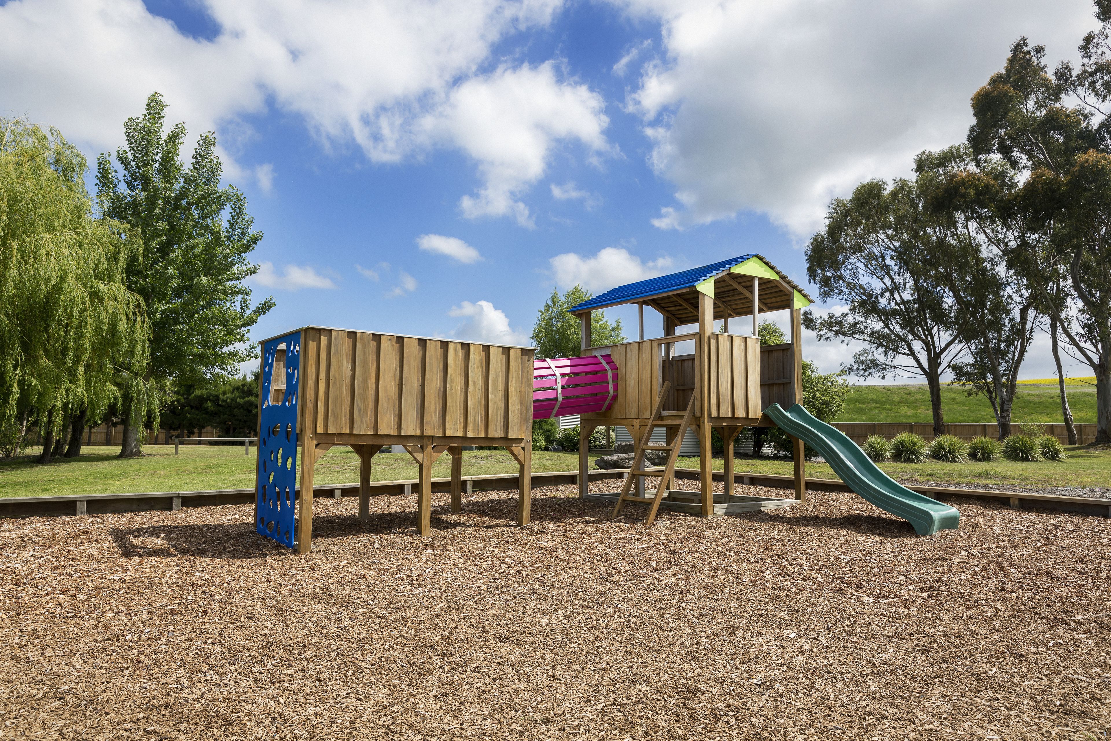 A wooden playground structure with a slide and a climbing area, surrounded by trees and a clear sky.
