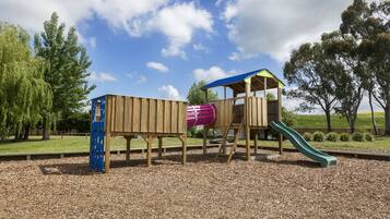 A wooden playground structure with a slide and a climbing area, surrounded by trees and a clear sky.