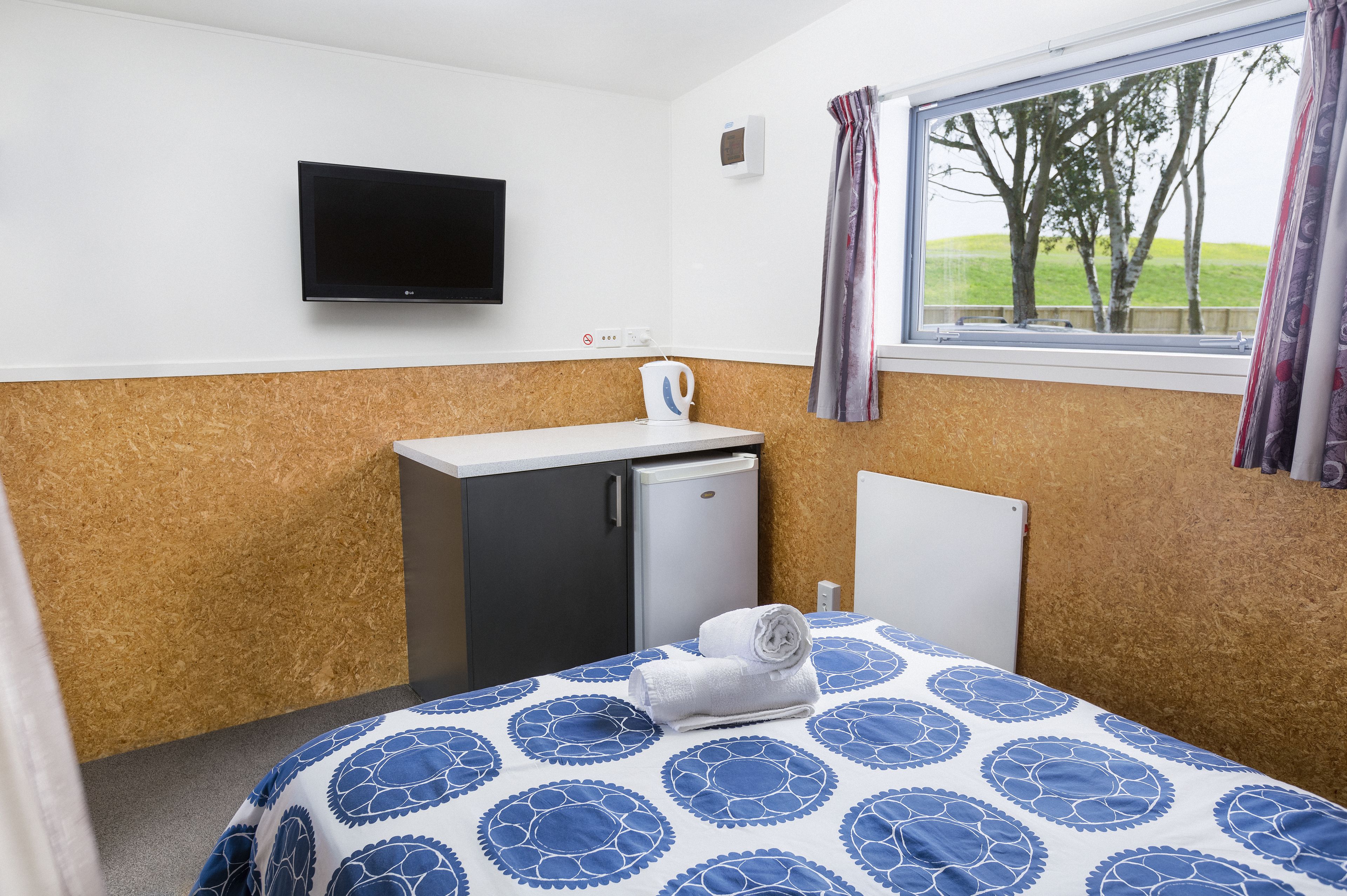 Habitación de hotel con cama, un escritorio pequeño con hervidor, televisor y ventana con vista a árboles.