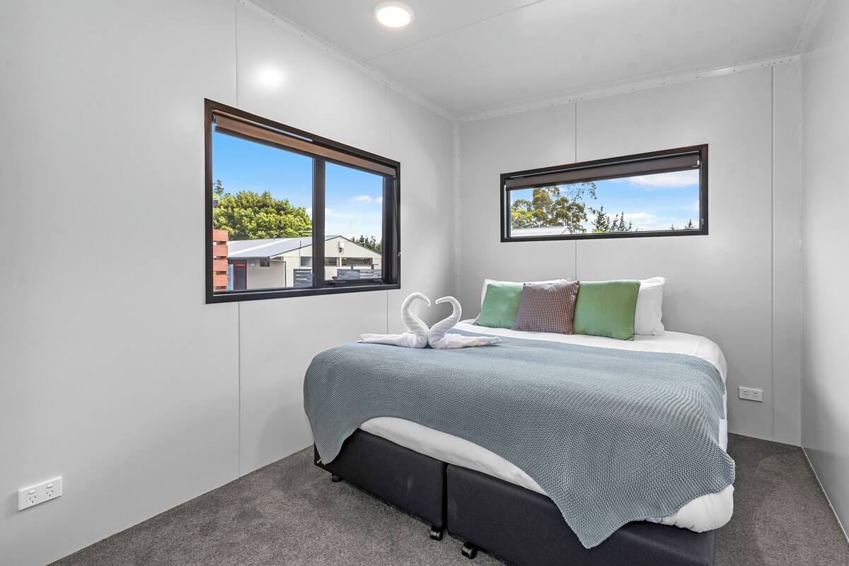 Ein modernes Schlafzimmer mit einem Bett, zwei Fenstern und Blick auf ein Haus und Bäume.