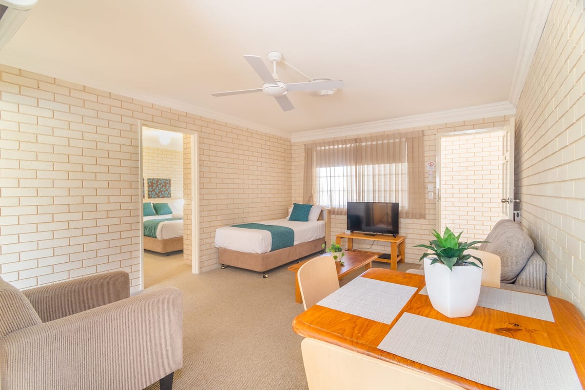 A hotel room with a brick wall, a bed, a TV, a dining table with a plant, and a ceiling fan.