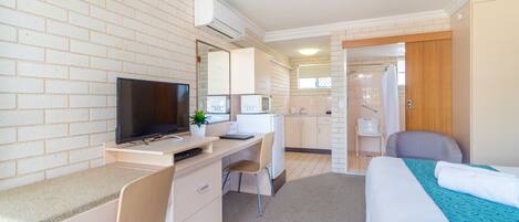 A hotel room with a bed, a desk with a TV, a chair, a small kitchenette, and a bathroom visible through an open door.