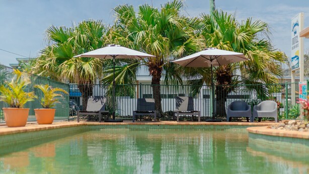 A pool area with lounge chairs and umbrellas, surrounded by palm trees and a fence. - Santa Fe Motel & Holiday Units (Lennox Head)
