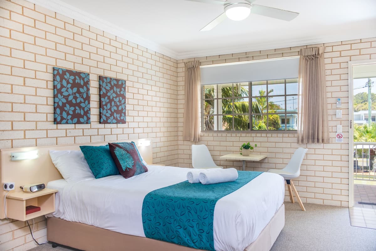 A hotel room with a brick wall, a bed with white and teal bedding, a ceiling fan, a window with curtains, and a small table with a chair.