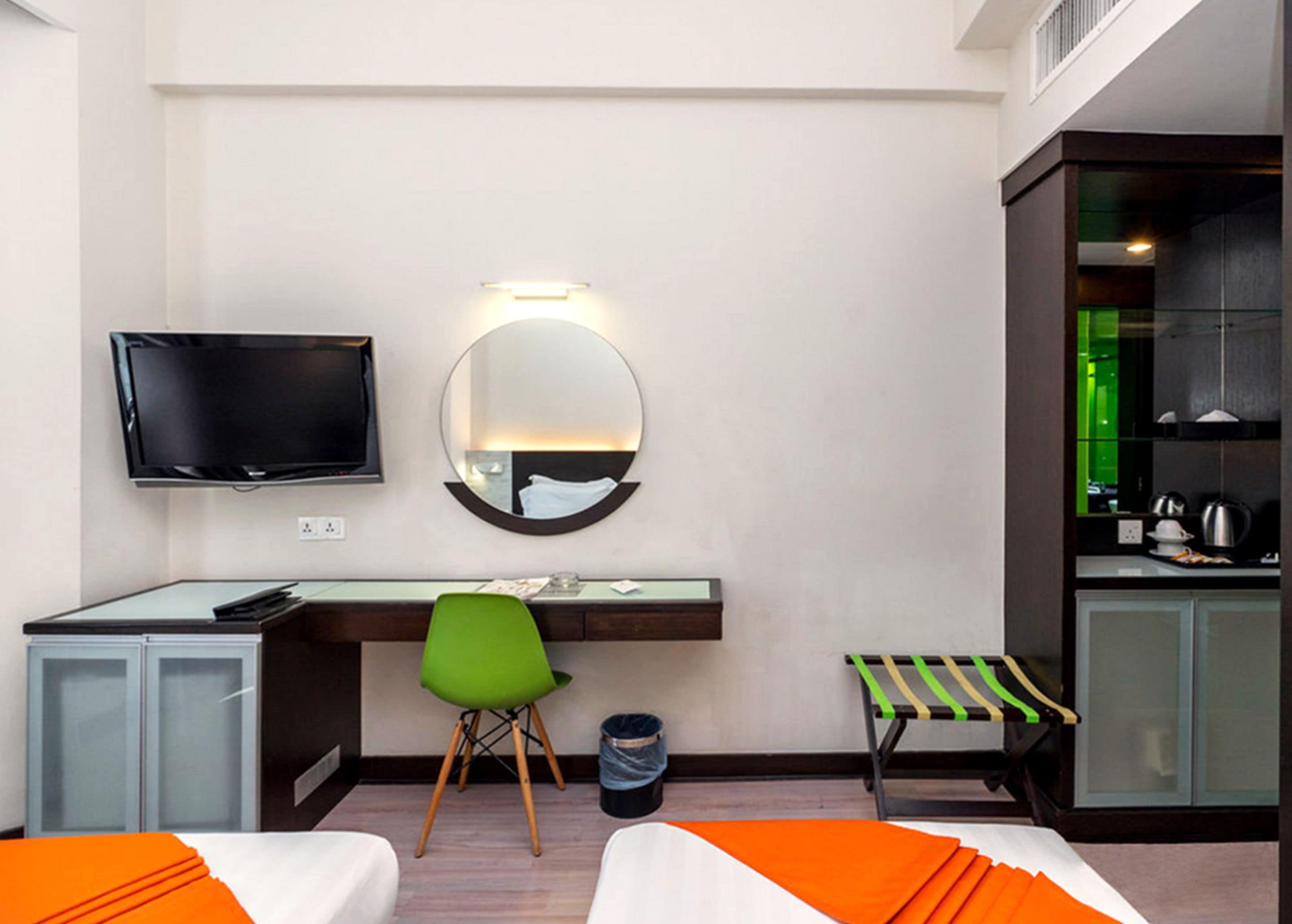 A hotel room with a bed, a desk with a chair, a TV, a mirror, and a kitchenette.