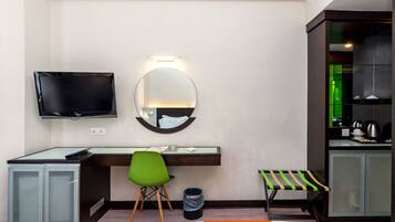 Una habitación de hotel con cama, escritorio con silla, televisión, espejo y kitchenette.