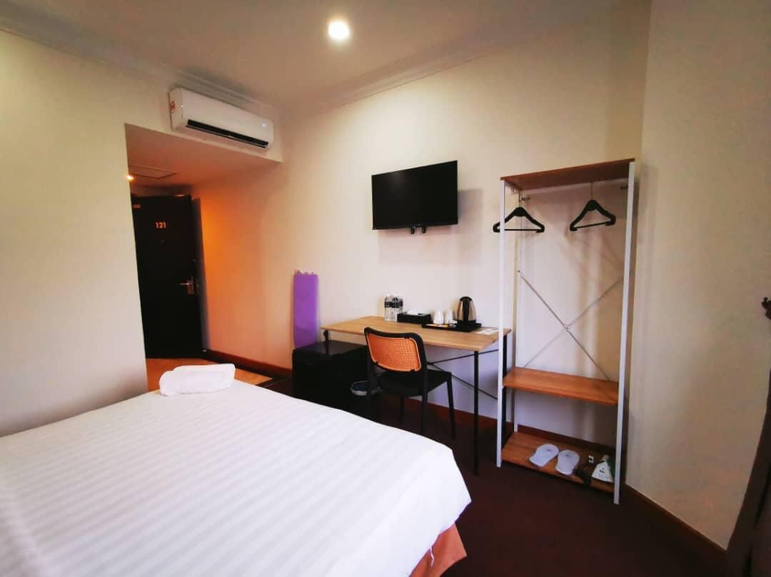 A hotel room with a bed, a desk with a chair, a TV mounted on the wall, and a coat rack with hangers.
