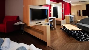 A modern hotel room with a TV, a small desk, a red sofa, a wooden partition, and a dining area with a bar.