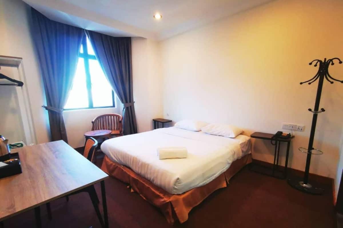 A hotel room with a bed, a desk, a chair, a coat rack, and a window with curtains.