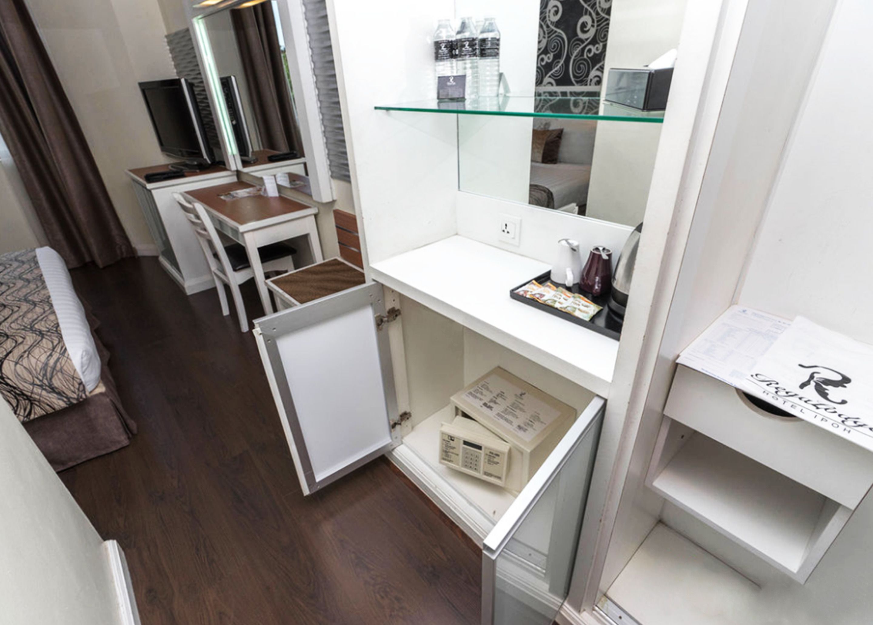 A compact hotel room with a kitchenette, a small desk, a mirror, and a glass shelf with toiletries.