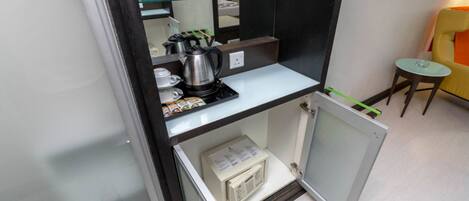 Una kitchenette funcional con cafetera, hervidor y microondas. Un estante de vidrio sostiene botellas y una pequeña mesa con una silla.