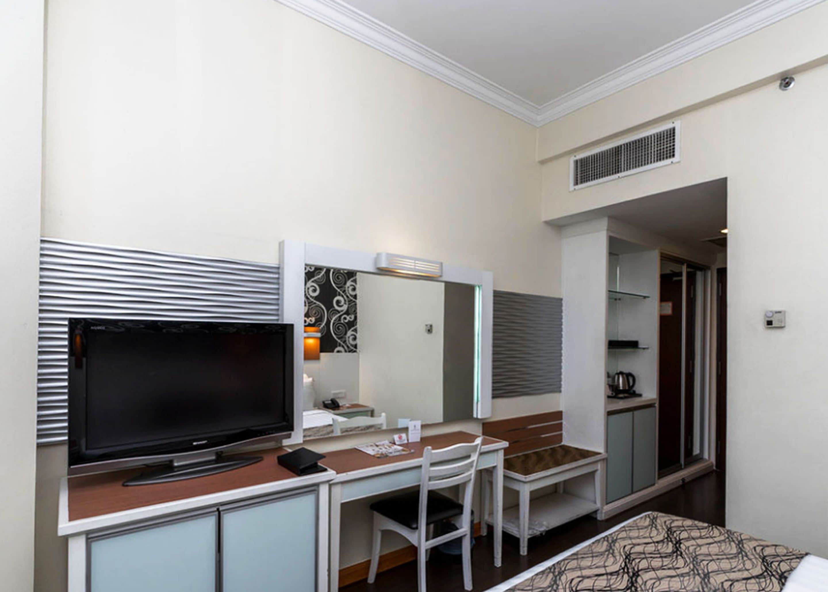 A hotel room with a TV, a desk, a chair, a bed, a mirror, and a wardrobe.