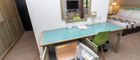 Une chambre d’hôtel de taille modeste, comprenant un lit simple, un bureau avec un miroir, une télévision et un petit réfrigérateur.