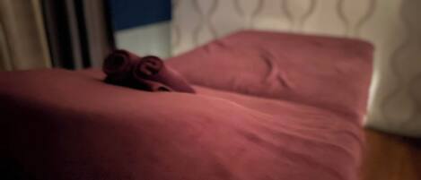 A room with a bed covered in a red blanket, a rolled-up towel on the bed, and a patterned wall in the background.